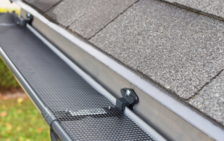 A close-up of a grey metal rain gutter installed on a roof with a black mesh gutter guard preventing leaves and debris from entering.