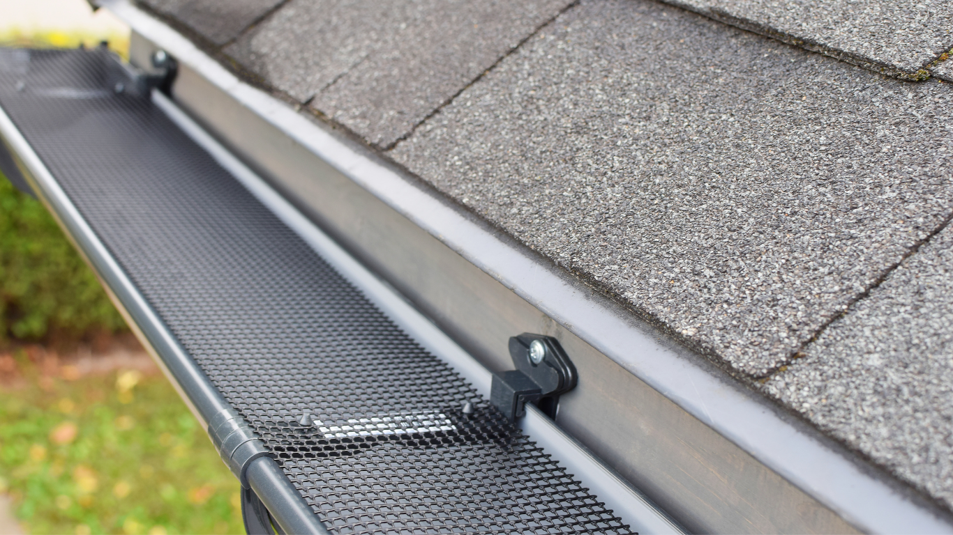 A close-up of a grey metal rain gutter installed on a roof with a black mesh gutter guard preventing leaves and debris from entering.